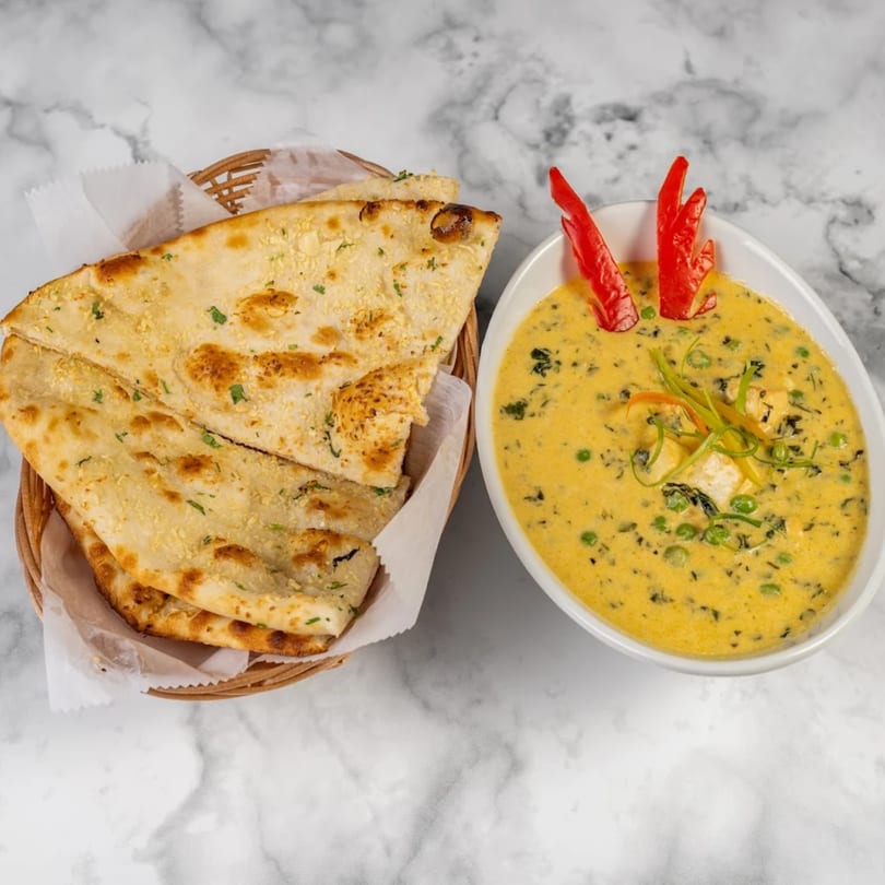Indian naan bread in a basket next to a bowl of creamy yellow curry with peas and paneer.