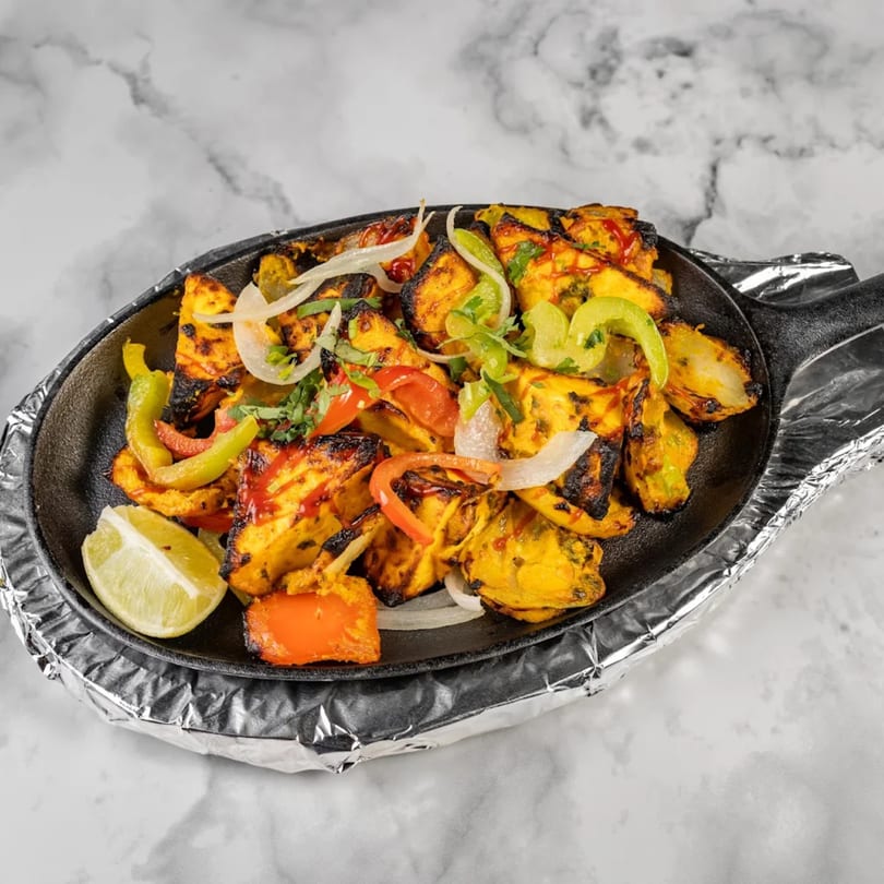 Indian paneer tikka, bell peppers, and onions in a sizzling cast iron pan with a lime wedge.