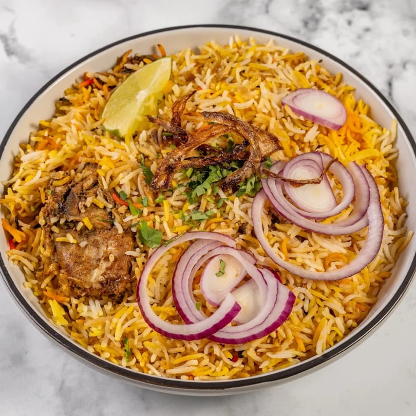 Indian chicken biryani with rice, fried onions, red onion slices, and a lime wedge in a bowl.